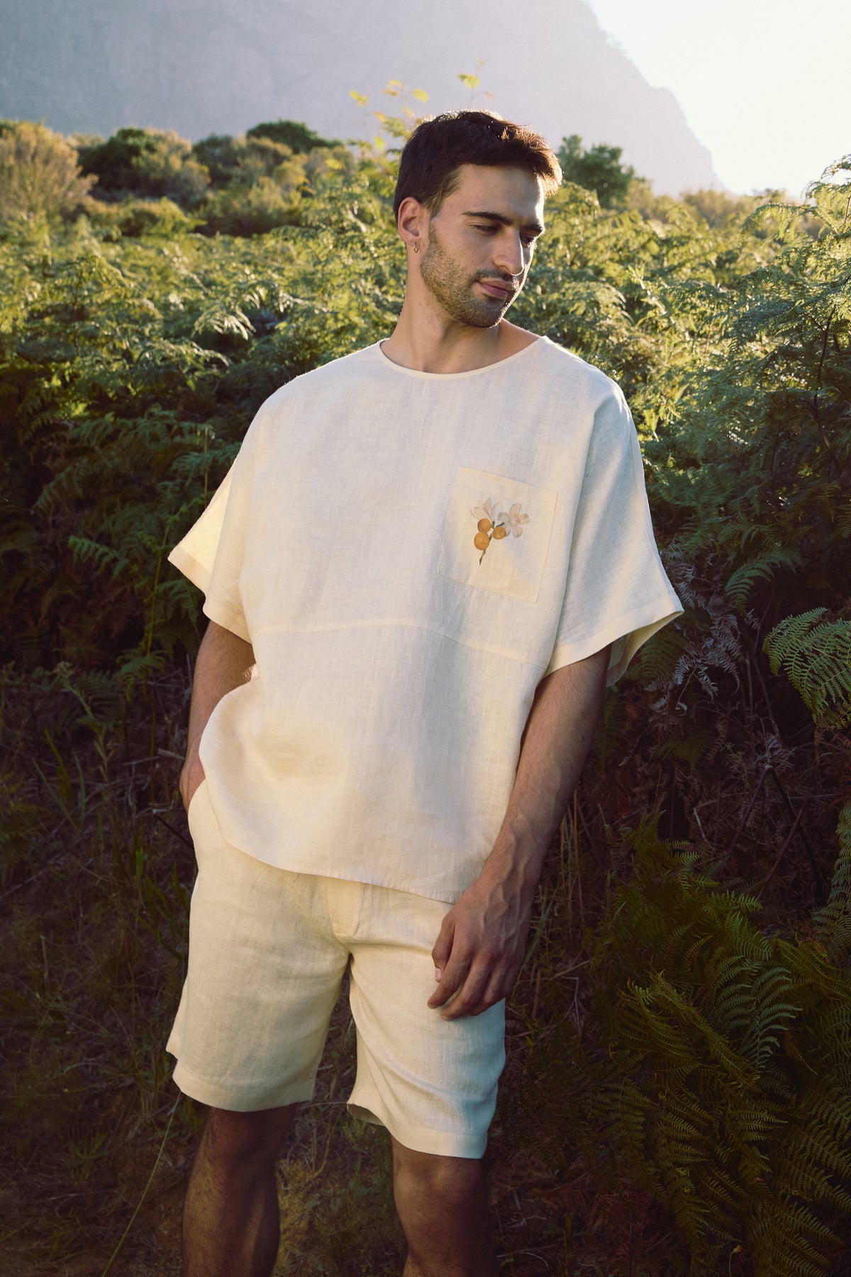 Man wearing a white oversized short-sleeve shirt with embroidered pocket and matching shorts in a garden